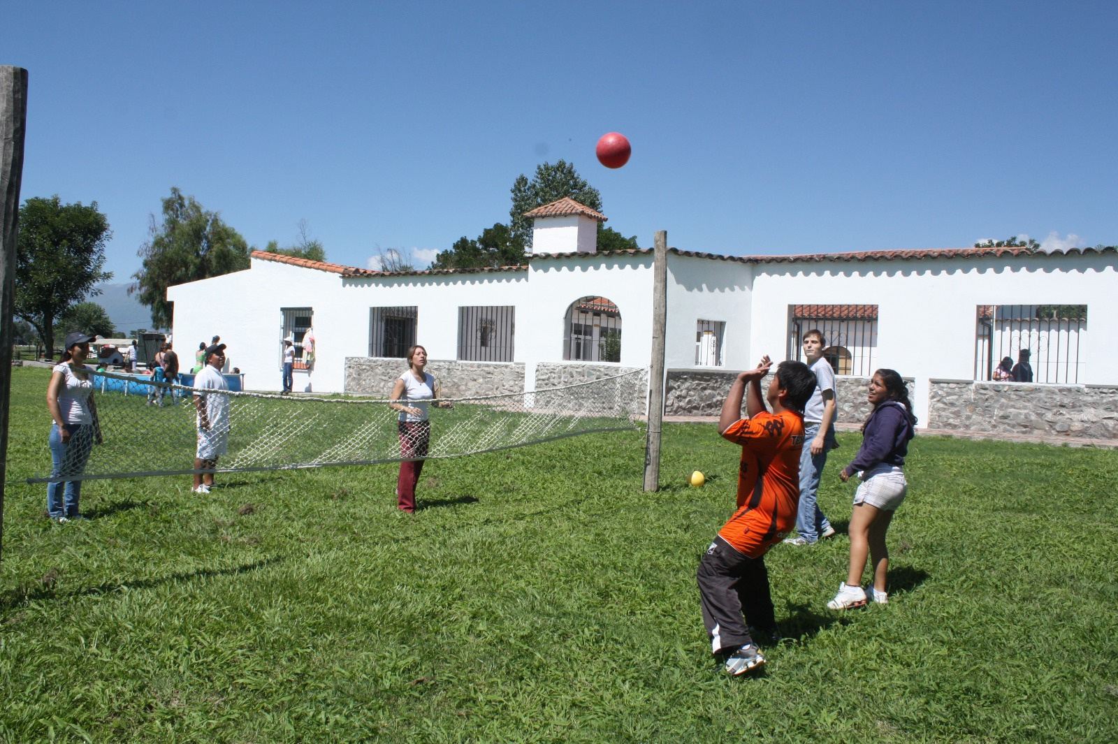 Jóvenes juegan con una pelota roja en el parque, frente a una casa blanca.