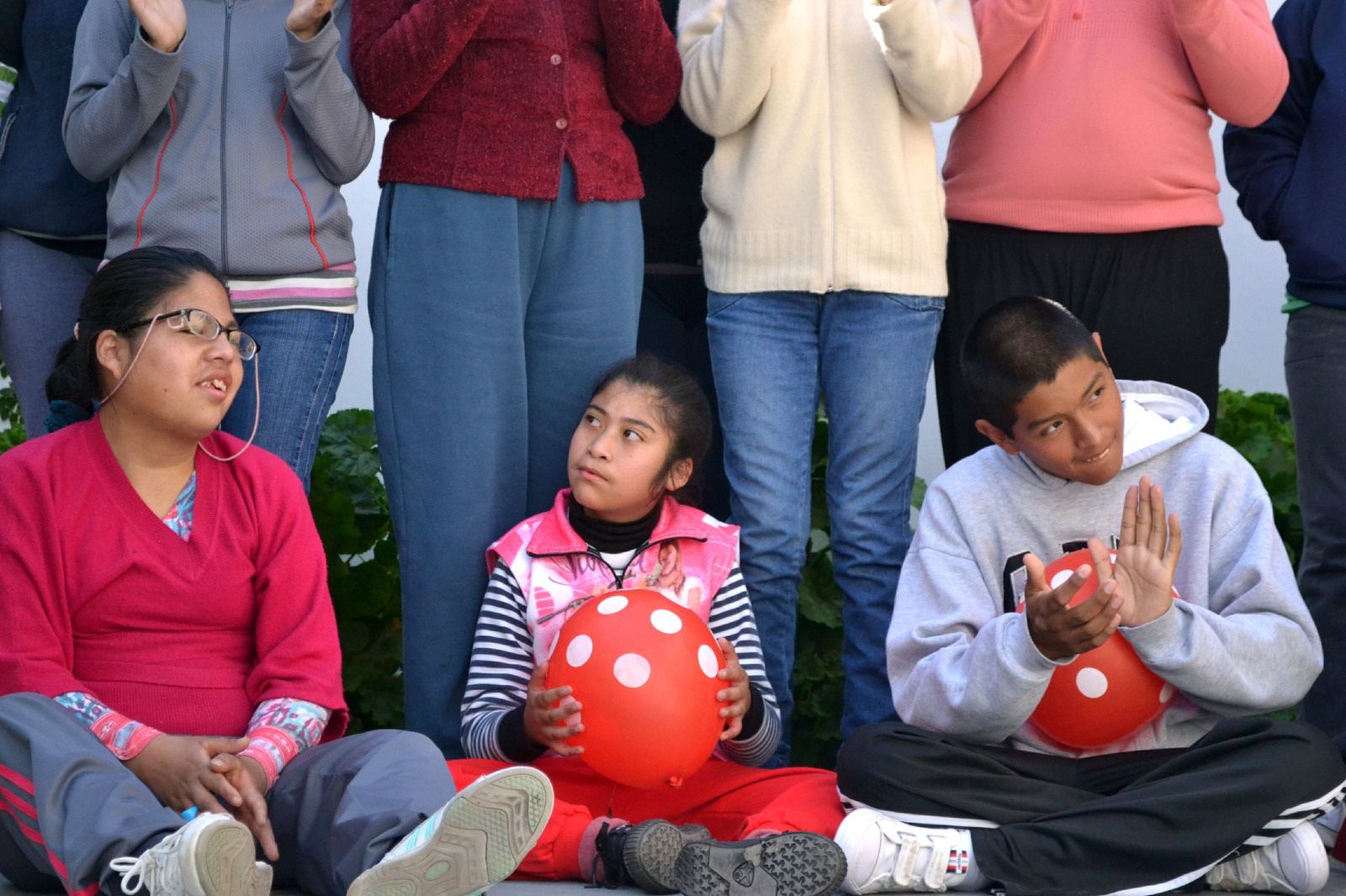 Jóvenes sentados en el pasto con pelotas rojas, junto a personas de pie.