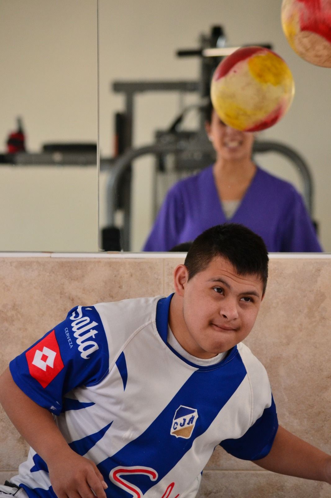 Un joven sonríe mientras juega con una pelota en el gimnasio; detrás, una profesional.